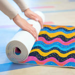 Person rolling up Carnival Currents yoga mat on studio floor, wavy multicolor stripes with sprinkles