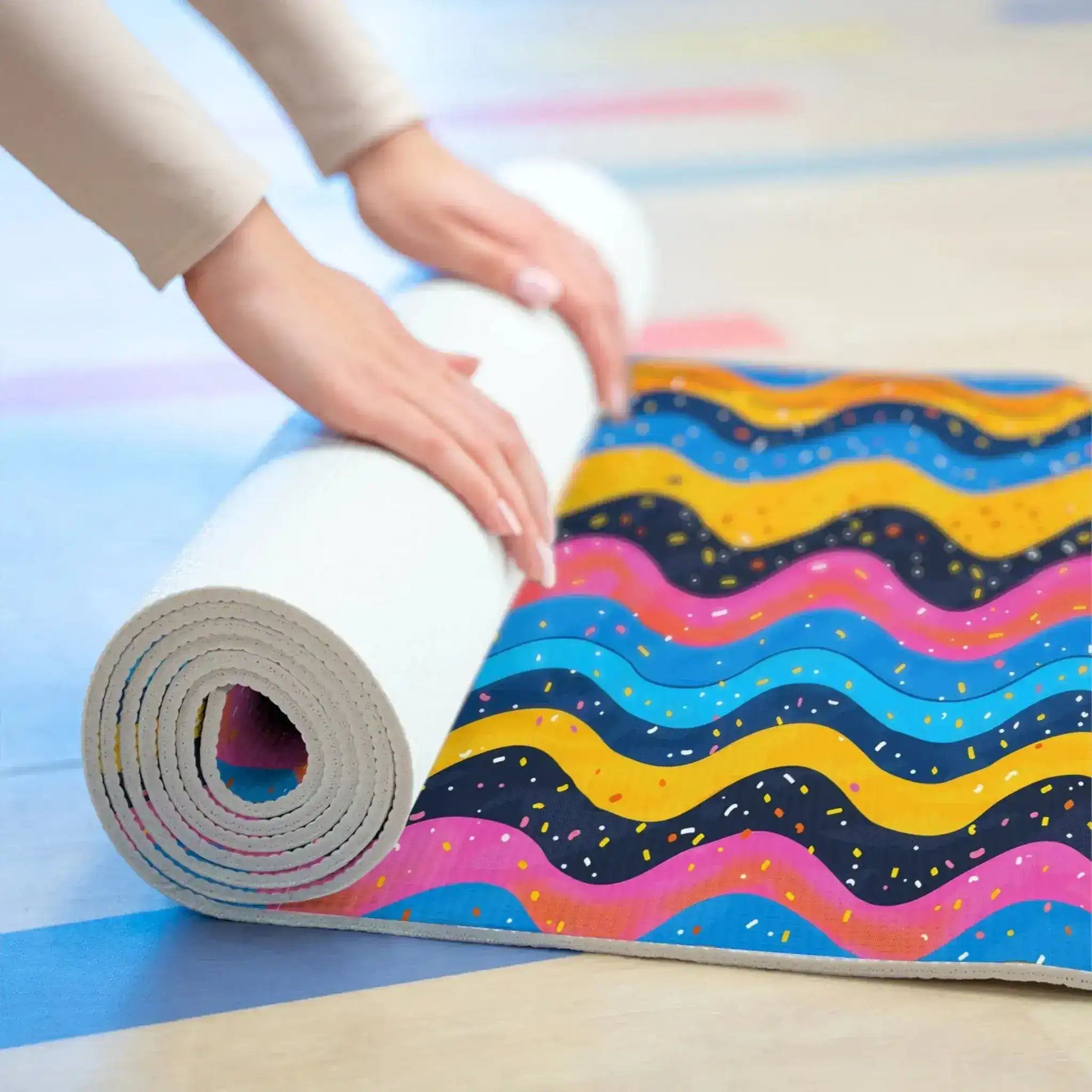 Person rolling up Carnival Currents yoga mat on studio floor, wavy multicolor stripes with sprinkles