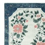 Corner view of the Petal Crest Bloom Hand Tufted Area Rug showcasing its elegant floral pattern. This exquisite wool rug, perfect for living rooms, features lush pink flowers and green leaves on a soft background, framed by an intricate blue border, making it a stunning choice for those seeking a floral rug to enhance their home decor.
