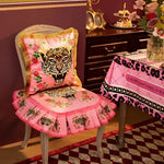 Pink leopard face throw pillow with gold fringe, featuring a vibrant design with floral accents, placed on a chair in a cozy interior setting.