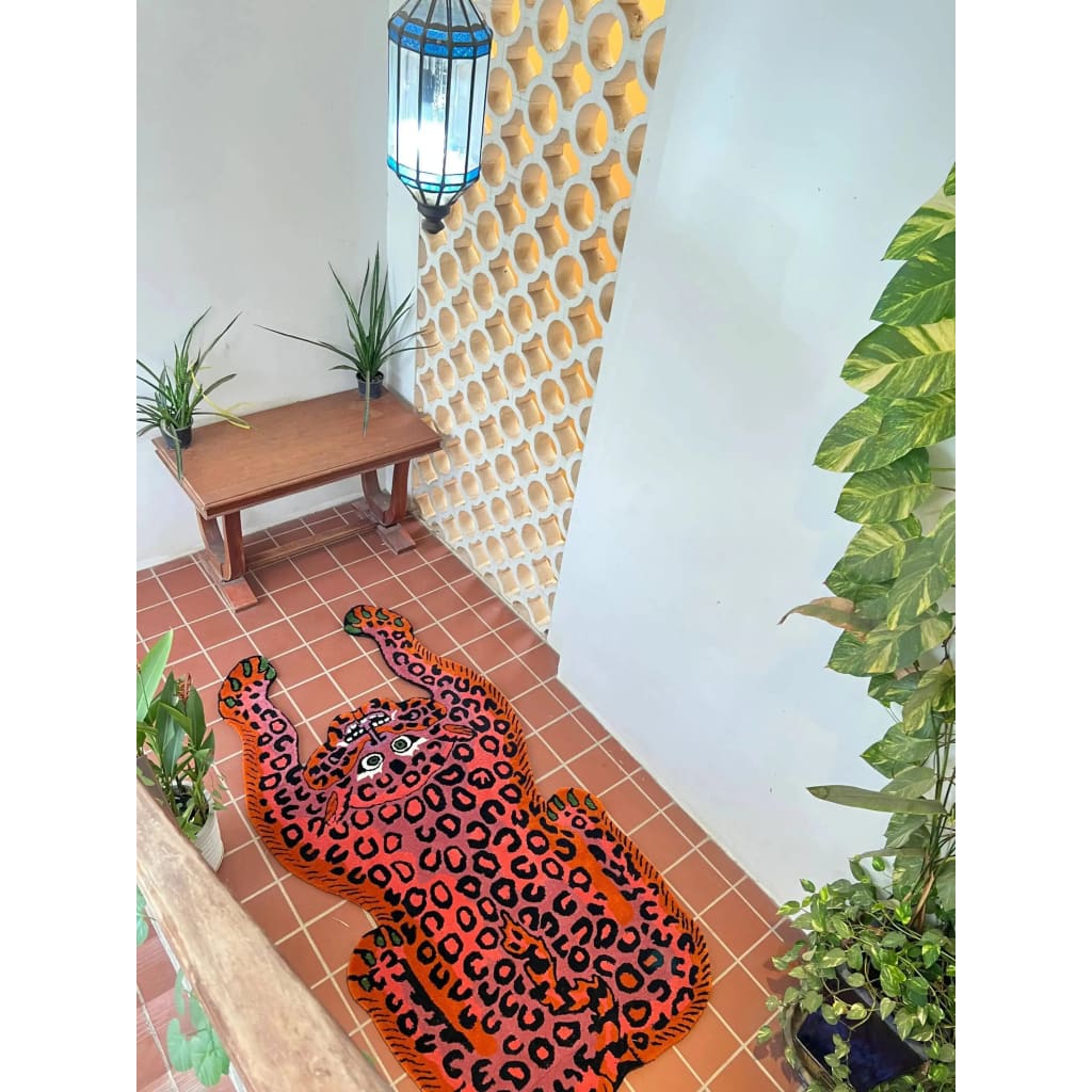 A vibrant Pink Leopard Yogi Accent Wool Rug adds a playful touch to a modern living space. The large area rug features a bold leopard design in striking pink and orange tones, perfect for those seeking unique living room rugs 8x10. Surrounded by plants and a wooden bench, this wool rug brings a vintage charm to the tiled floor, ideal for enhancing any living room decor.