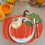 Orange pumpkin-shaped placemat with ribbed segments and tan border, pumpkin plate and autumn props on table