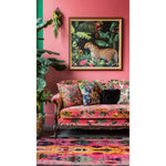 A vibrant living room setting featuring a radiant spectrum wool hand-knotted area rug. The square area rug, with its intricate abstract patterns in bold colors, complements the floral-themed sofa and lush indoor plants. The wool rug adds a touch of elegance and warmth to the living room decor.