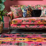 A vibrant Radiant Spectrum Wool Hand Knotted Area Rug with colorful abstract patterns, placed elegantly in a stylish living room setting. The square rug complements the floral-patterned pink sofa, adding a pop of color and texture to the space. Ideal for those seeking unique living room area rugs that offer both comfort and a splash of artistic flair.