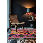 A vibrant square area rug made of wool, featuring a colorful abstract pattern, is placed in a stylish living room. The rug adds a splash of color to the dark-toned space, complementing the wicker chair and elegant side table with a lamp and vase of flowers. Perfect for enhancing modern living room aesthetics.