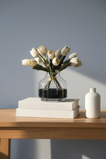 Real touch artificial tulips in a clear glass vase on stacked books against a soft blue wall.