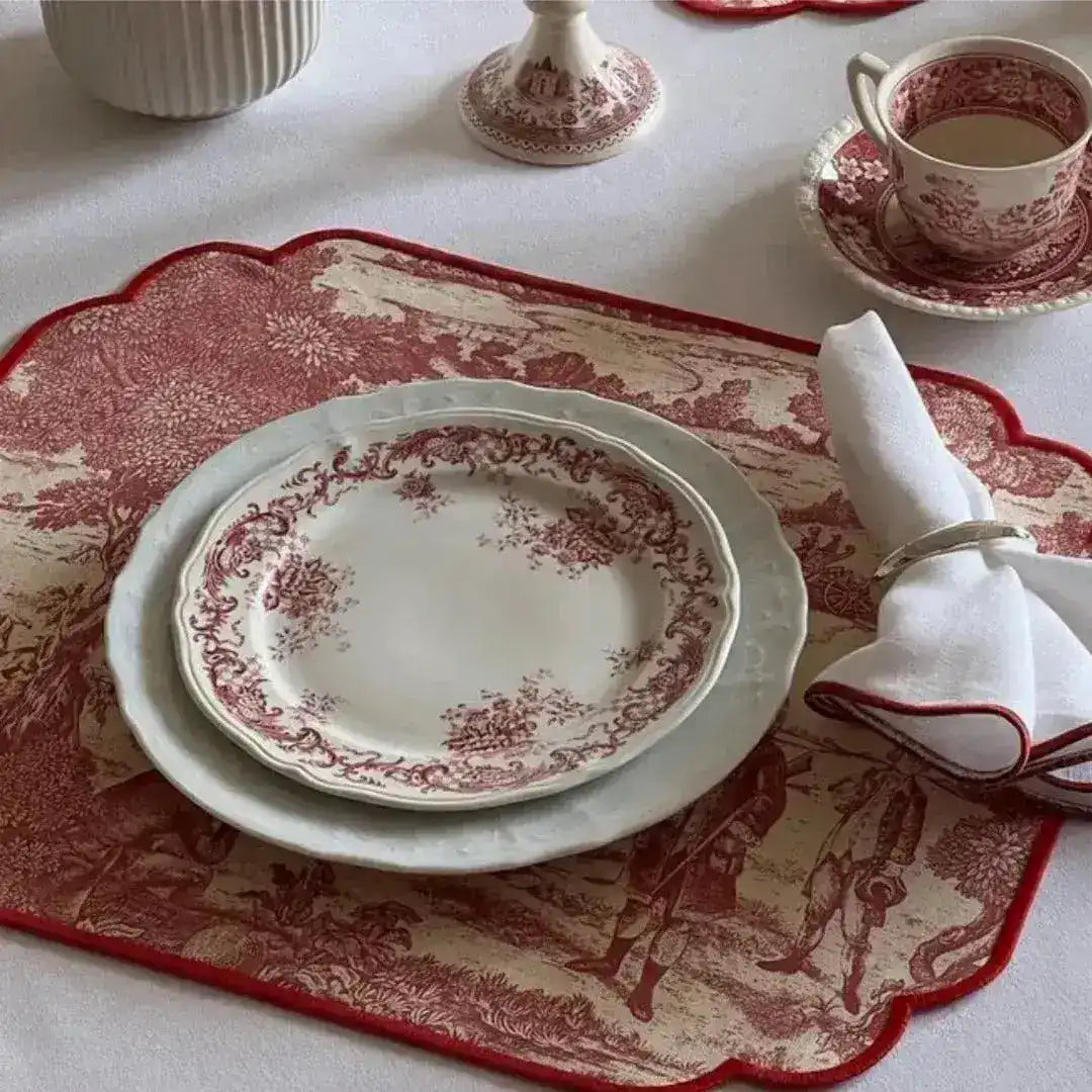 red and cream placemats with scalloped border and toile pastoral print, styled with stacked china plates and rolled white napkin