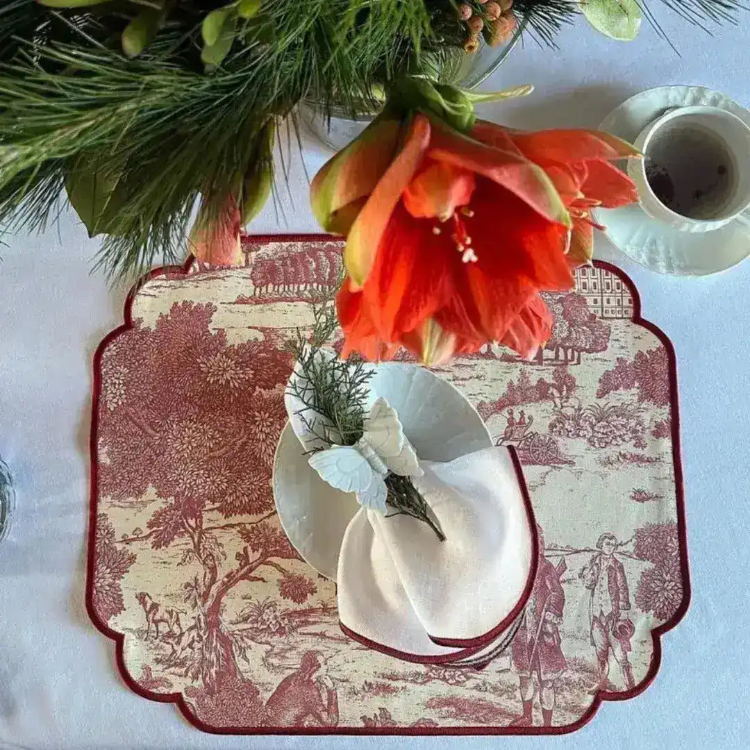 red and cream placemats overhead view with toile print, white napkin and butterfly ring, orange amaryllis and pine greenery