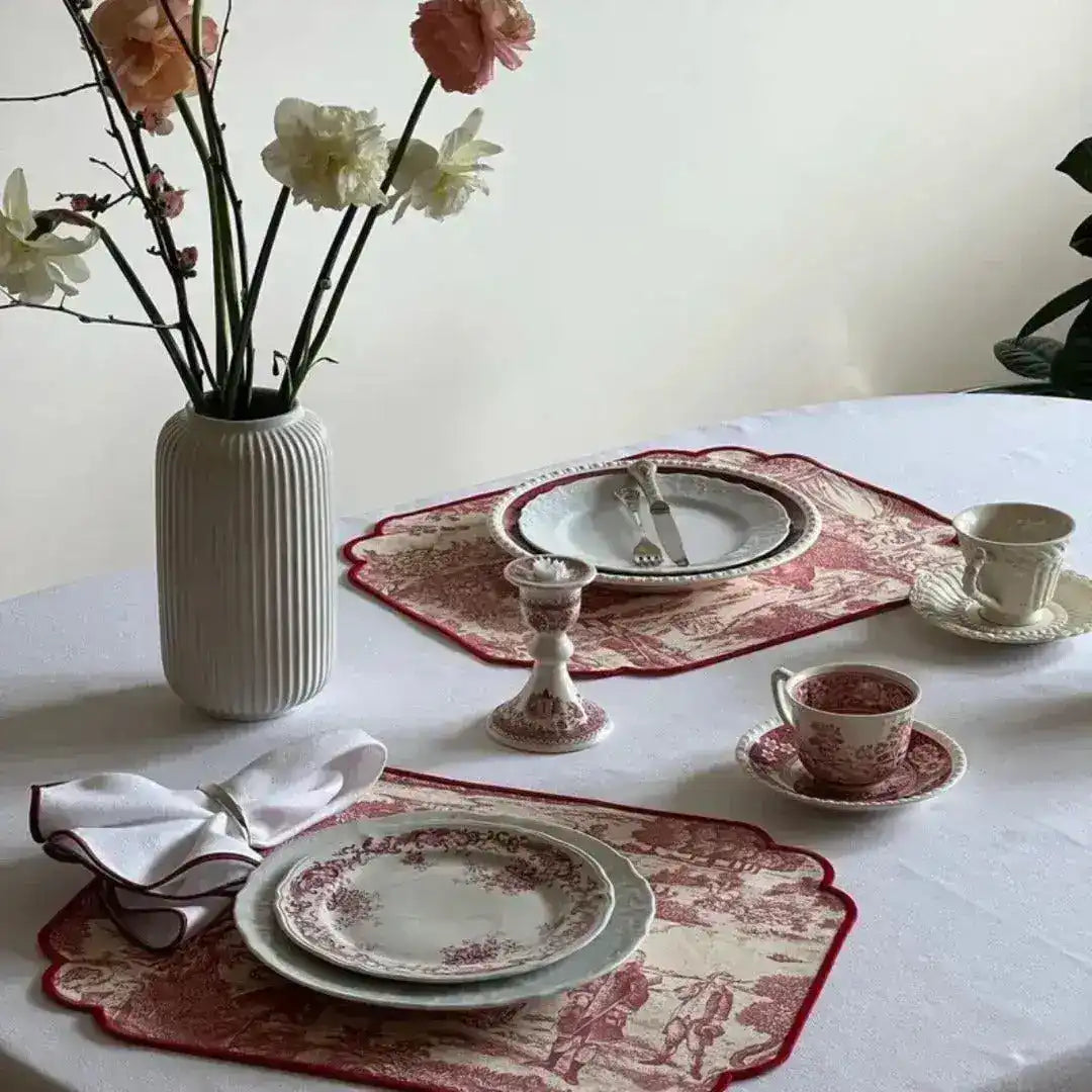 two red and cream placemats with toile print on white table with floral china, white vase with pink peonies and candleholder