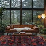 A beautifully decorated living room featuring a Red Bloom Harmony area rug. The hand-tufted wool rug showcases a vibrant floral design, perfect for adding a vintage touch to your living room or dining room. This 8x10 rug complements the warm tones of a leather sofa and the serene view of a large window overlooking lush greenery.