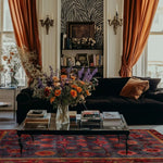 A beautifully styled living room featuring the Red Bloom Harmony Rug, an exquisite hand-tufted area rug designed with a floral pattern. This 8x10 vintage wool rug adds a touch of elegance and warmth to the space, making it ideal for dining room areas as well. The rug's vibrant red hues complement the rich decor, including a plush black sofa, a glass coffee table stacked with books, and an array of colorful flowers in a vase, set against luxurious orange curtains and a zebra-patterned wall.