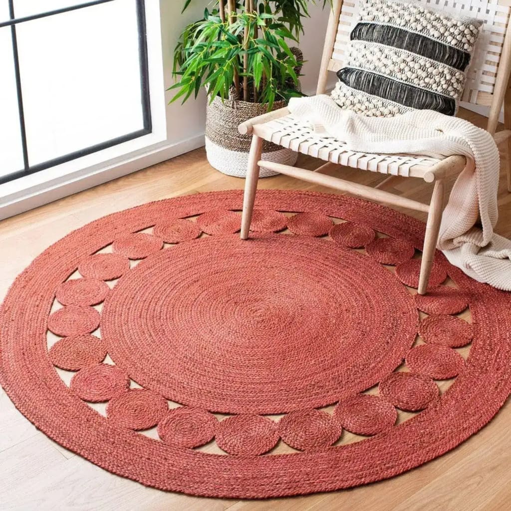 A cozy living room setting featuring a red round jute area rug. The large area rug, perfect for living rooms, adds a warm touch to the wooden floor. The intricate patterns on the round rug enhance the room's aesthetics, complemented by a wooden chair with a textured cushion and a soft throw blanket.