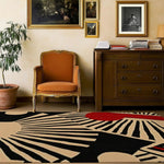 A stylish living room setting featuring a Rhythmic Rays Hand Tufted Area Rug. The rug showcases an abstract design with bold black, beige, and red patterns, perfect for enhancing the aesthetics of any living room or dining room. This wool rug complements the classic decor, including an orange upholstered chair, a wooden dresser, and framed artwork on the wall.