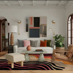 A stylish contemporary area rug with swirling patterns in black, pink, and orange hues, enhancing a modern office setting. The rug is placed under a chic glass-top coffee table, surrounded by a white sofa, colorful cushions, and elegant decor, making it an ideal wool rug for office spaces.
