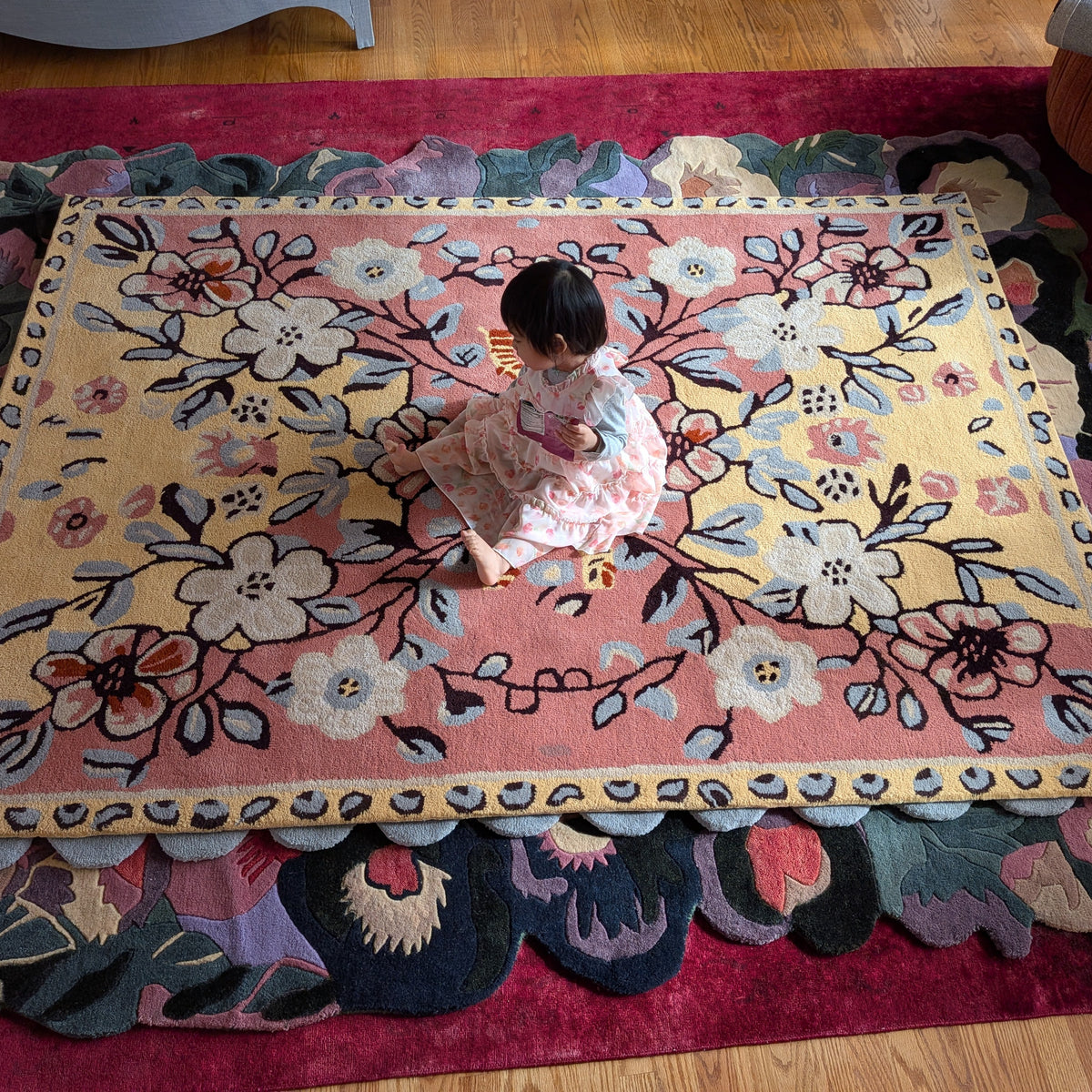 Child sitting on a floral-patterned rug in a room with wooden flooring.