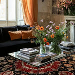 A cozy living room setting featuring a luxurious Royal Persian Hand Knotted Area Rug. This vintage rug, with its intricate patterns and deep red tones, complements the elegant decor, including a black sofa, orange cushions, and a glass coffee table adorned with fresh flowers and books. Ideal for dining room and living room spaces, this square wool rug adds a touch of sophistication and warmth.