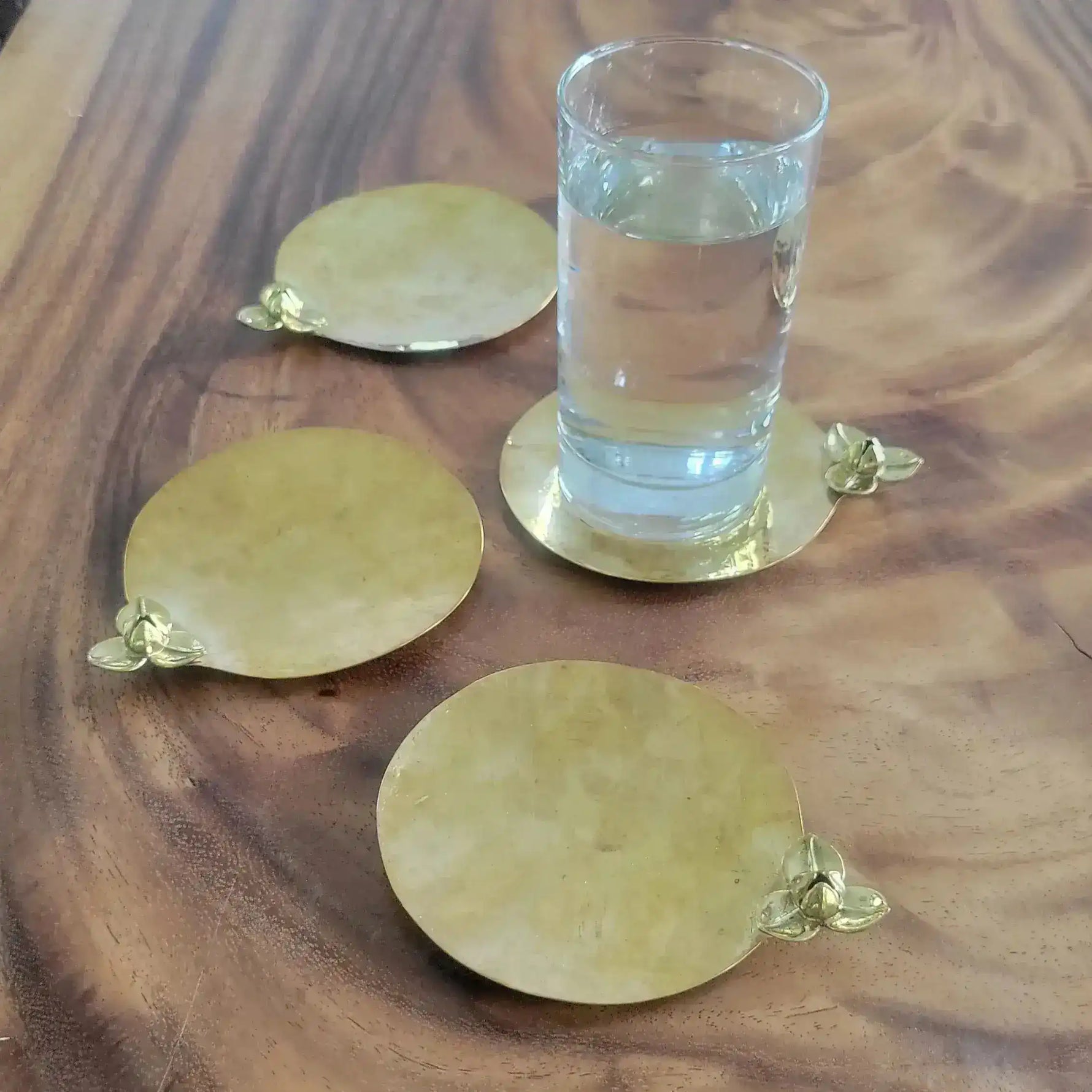 Set of four round gold brass Rumduol flower coasters on wood-grain tray, one supporting a tall water glass, showing 3D blossom detail