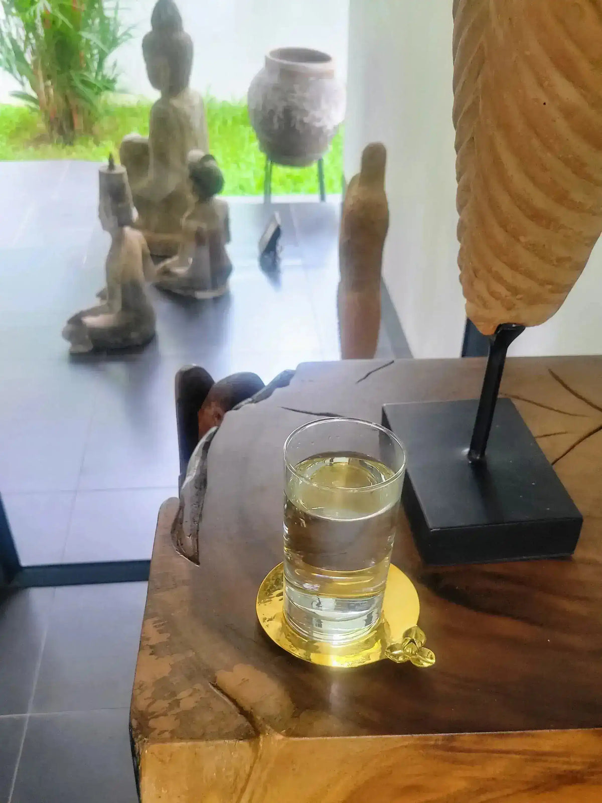 Single brass Rumduol flower coaster holding a glass of water on a live-edge wood table with Buddha statues and decor in background