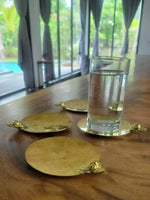 Three brass Rumduol flower coasters on a wood dining table, one holding a clear water glass, natural light from window behind