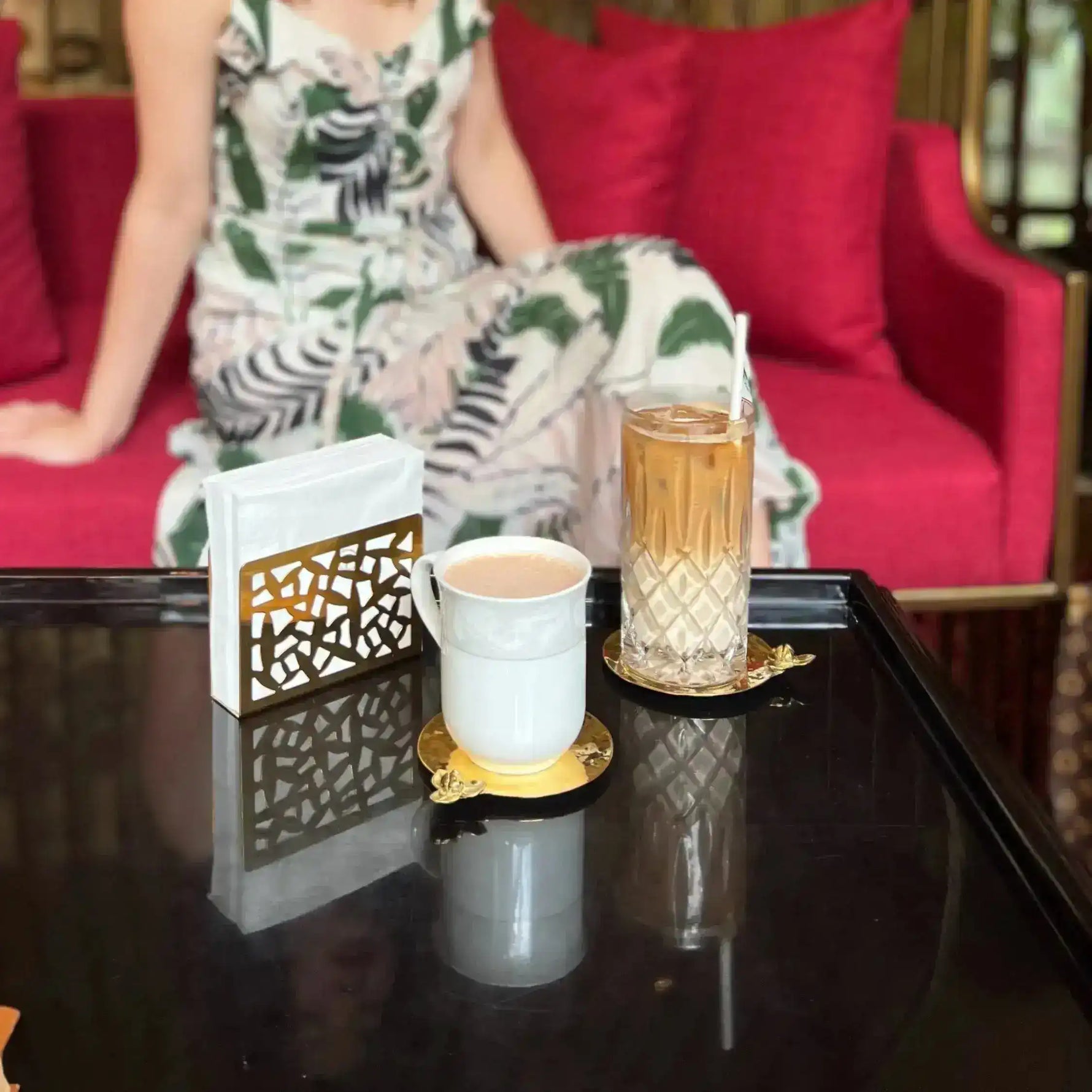 Two brass Rumduol flower coasters on a glossy dark table with a white mug and tall crystal glass, person on red sofa in background