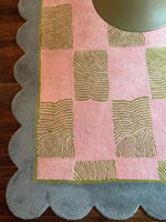 A close-up view of a Scalloped Leaf Checker Pastel Hand Tufted Area Rug, ideal for large living rooms. This wool rug features a pink and green checker design with a scalloped blue edge, perfect for adding a boho touch to your space. Its unique pattern and soft texture make it a standout among large area rugs for living rooms.