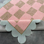 Pink and gold checkered rug with scalloped edges on a gray surface