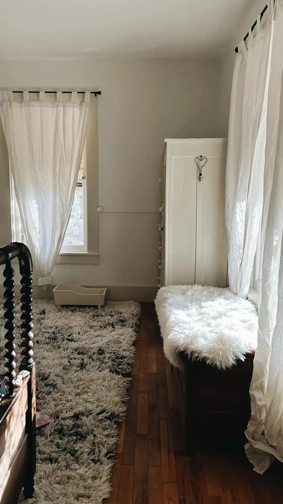 White sheepskin pelt rug on dark wooden bench in moody bedroom with spindle bed, gray area rug, sheer curtains