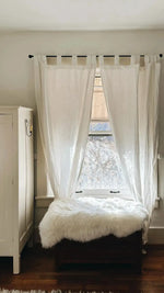 Sheepskin pelt rug draped over dark wooden bench by bright window with white sheer curtains and hardwood floor