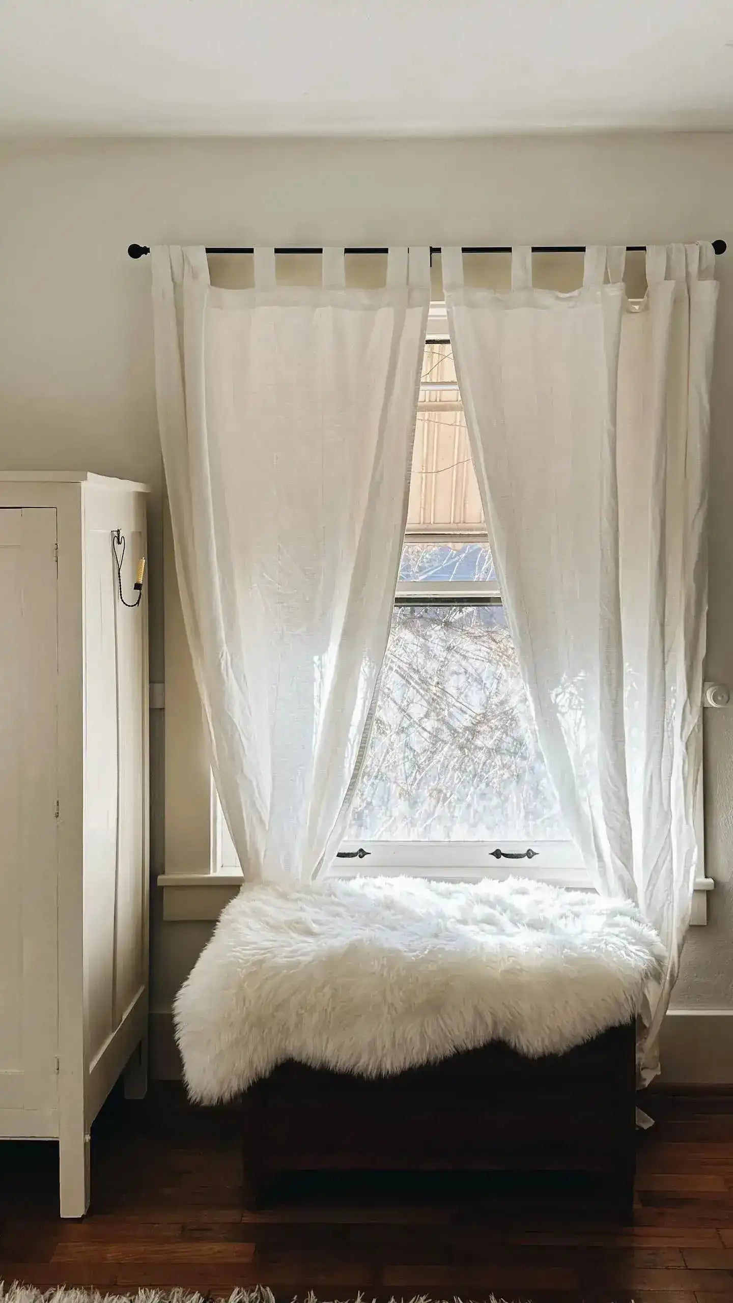Sheepskin pelt rug draped over dark wooden bench by bright window with white sheer curtains and hardwood floor