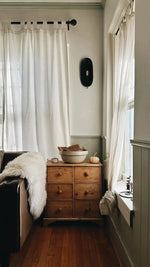 Fluffy sheepskin pelt casually draped over dark sofa arm beside pine dresser, white sheer curtains, wood floor