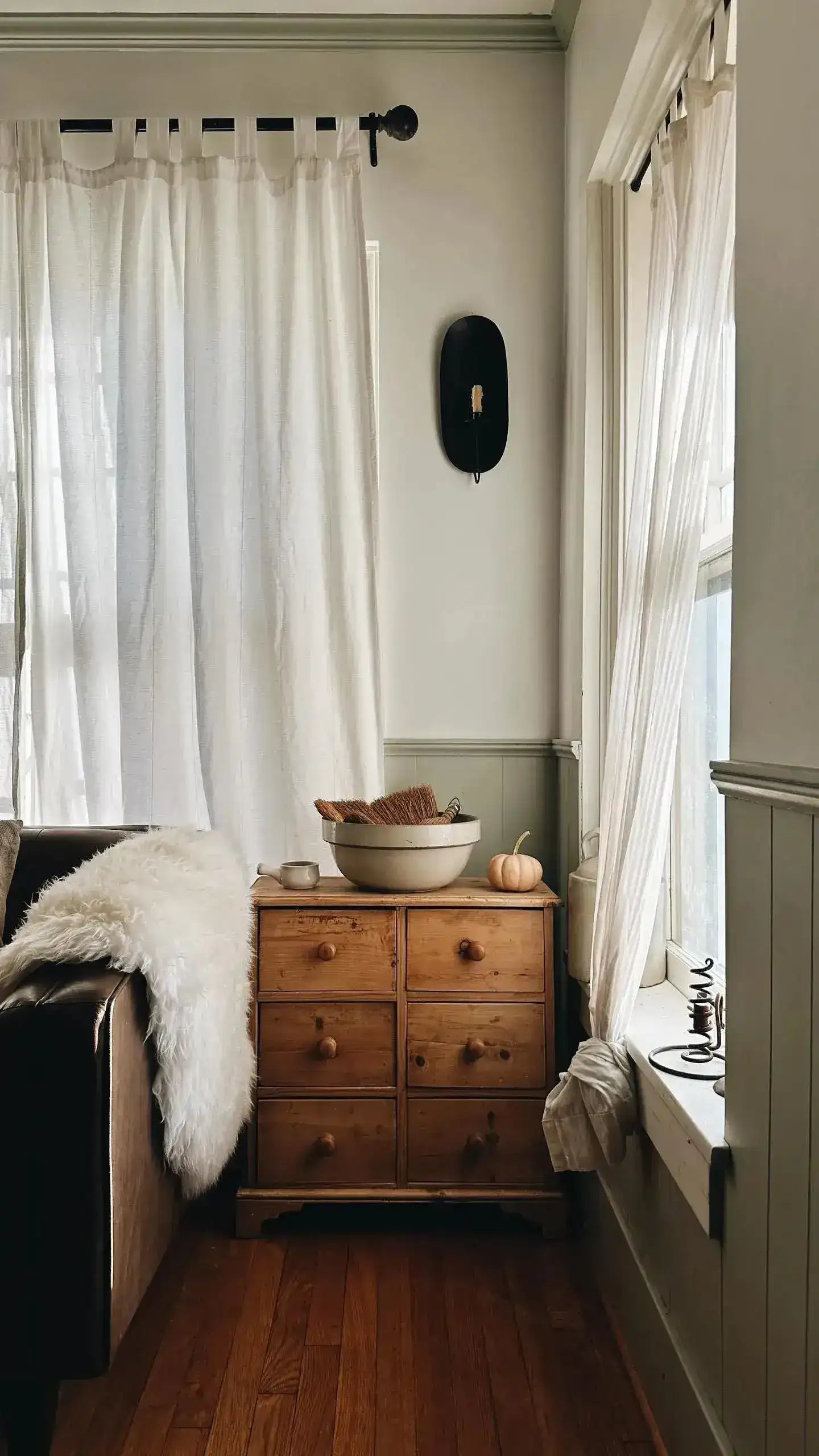 Fluffy sheepskin pelt casually draped over dark sofa arm beside pine dresser, white sheer curtains, wood floor