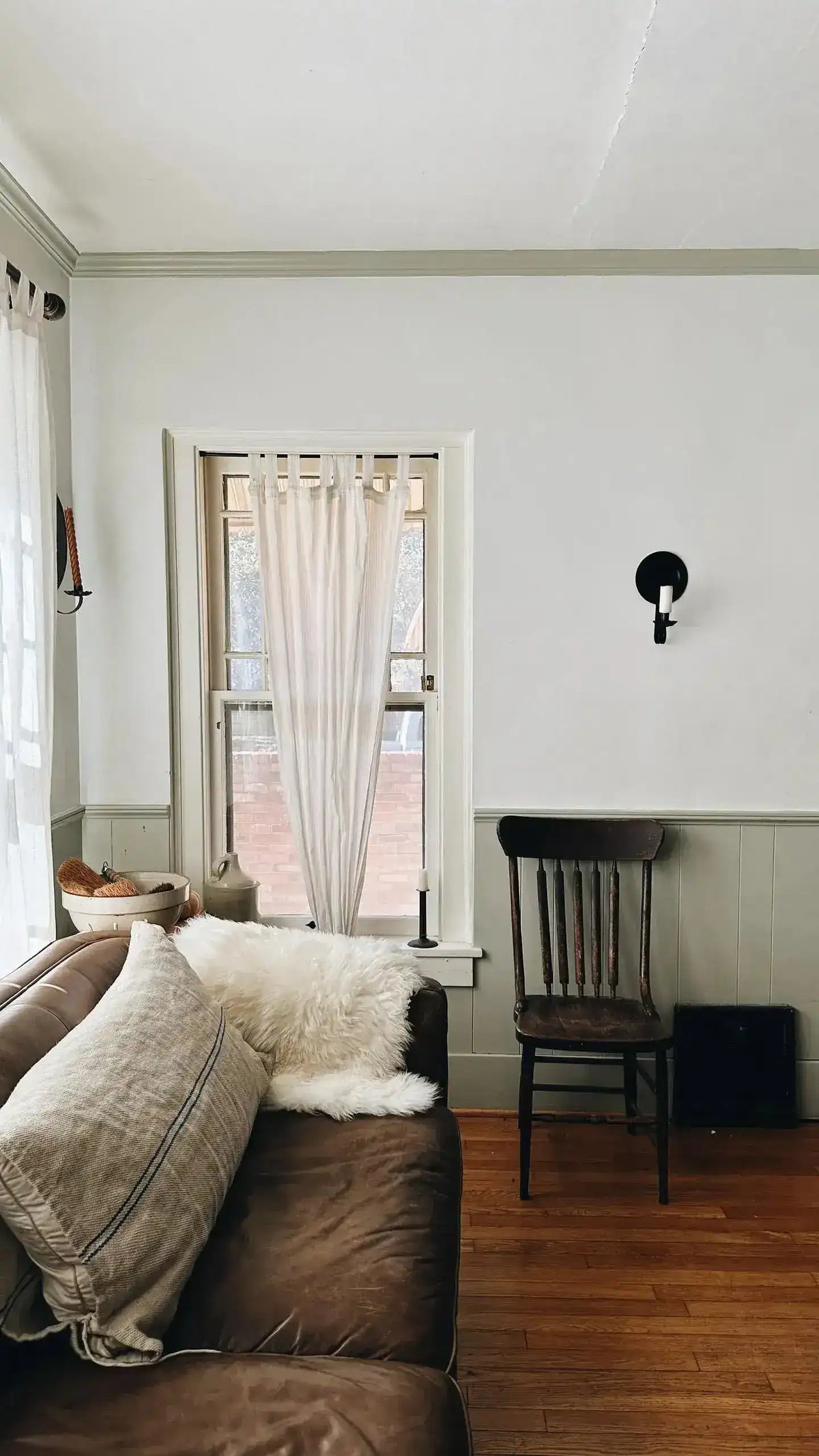 White sheepskin pelt rug tossed over brown leather sofa back in living room with wooden chair and curtained window
