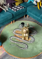 Top view of a stylish round wool rug in a dining room setting, featuring an intricate snake and apple design on a green backdrop. This elegant area rug complements the surrounding teal furniture, making it an ideal choice for formal dining room decor.