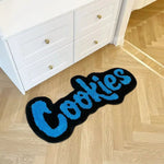 A quirky and playful area rug featuring the word 'Cookies' in bold blue letters on a black background, placed on a light wooden floor next to a white dresser. This handmade plush rug is a unique addition to any large living room rugs collection, adding a touch of fun and coziness.