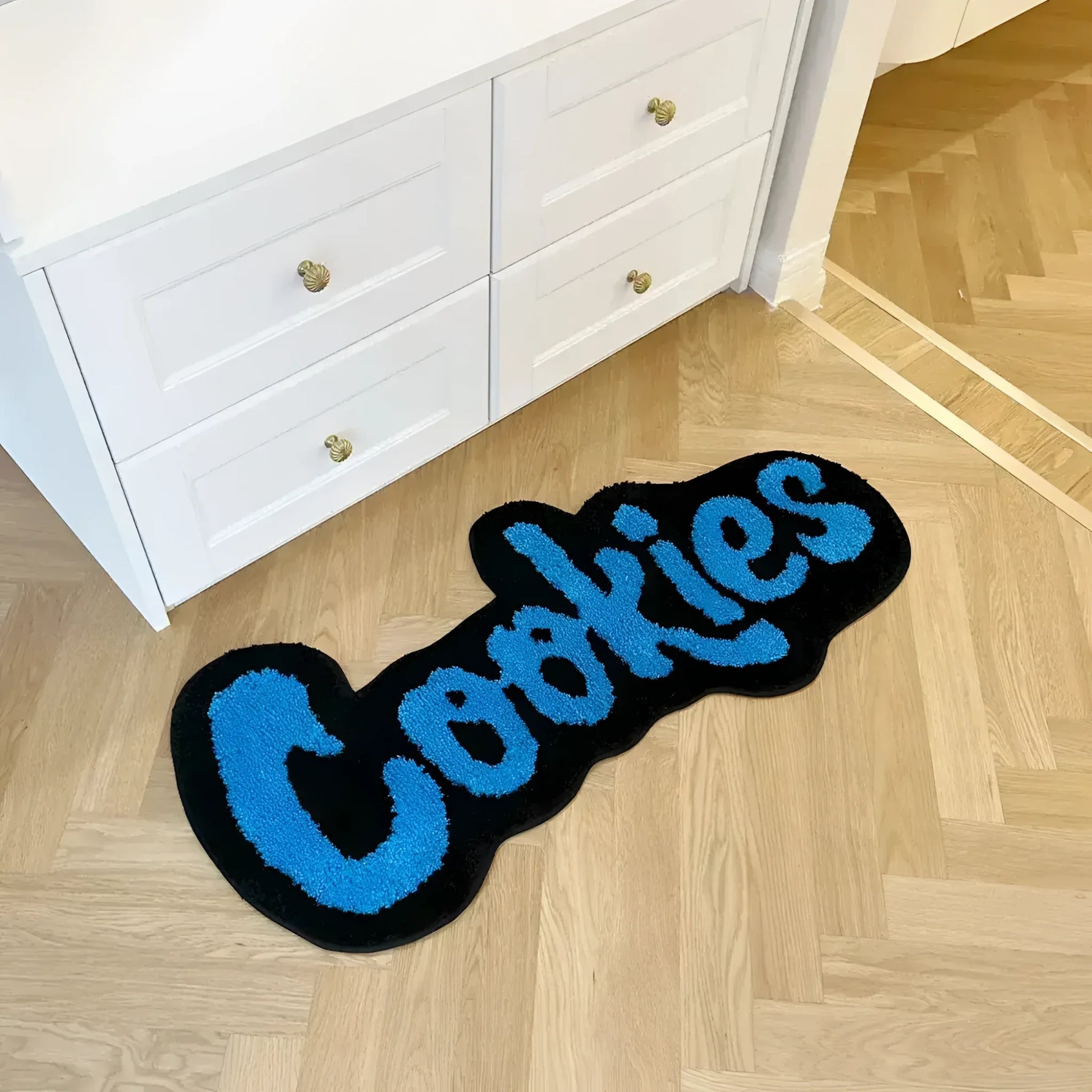 A quirky and playful area rug featuring the word 'Cookies' in bold blue letters on a black background, placed on a light wooden floor next to a white dresser. This handmade plush rug is a unique addition to any large living room rugs collection, adding a touch of fun and coziness.