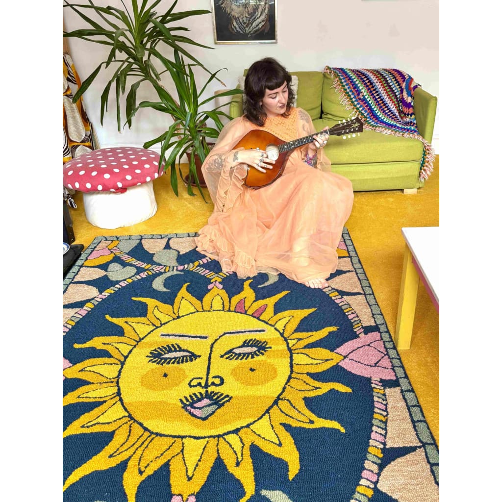 A vibrant area rug featuring a bold sun design in yellow and blue, perfect for adding a touch of whimsy to any living room. The wool rug is laid out in a cozy space with a woman playing a string instrument, surrounded by eclectic decor.
