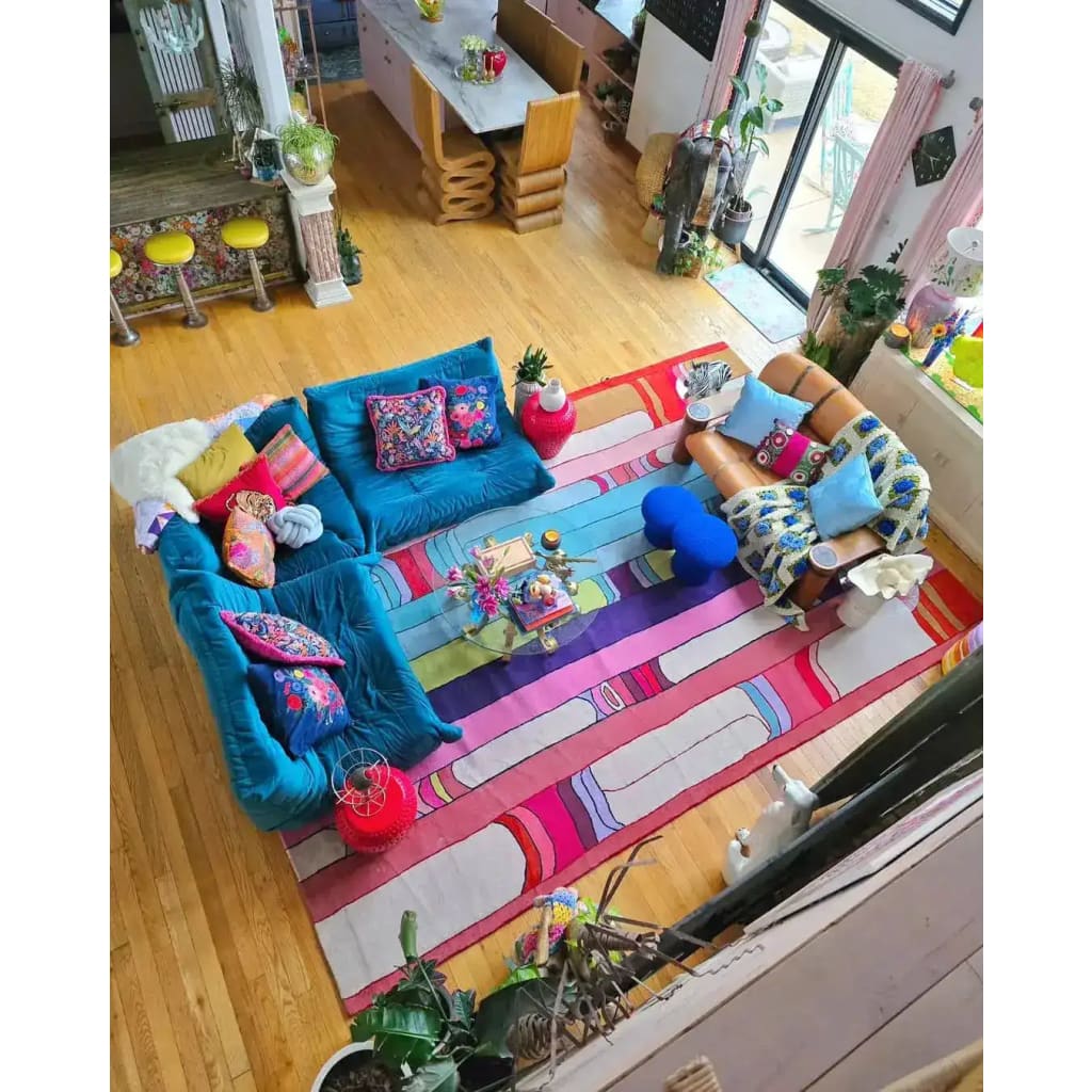 A vibrant and colorful Spectrum Stripe Hand Tufted Area Rug adorns a spacious living room with a contemporary design. The rug features bold, multicolored stripes that add a lively touch to the wooden floor. This wool rug, ideal for large living rooms, measures 8x10 and complements the eclectic, cozy seating arrangement, including a teal sofa and patterned cushions.