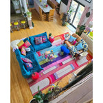 A vibrant and colorful Spectrum Stripe Hand Tufted Area Rug adorns a spacious living room with a contemporary design. The rug features bold, multicolored stripes that add a lively touch to the wooden floor. This wool rug, ideal for large living rooms, measures 8x10 and complements the eclectic, cozy seating arrangement, including a teal sofa and patterned cushions.