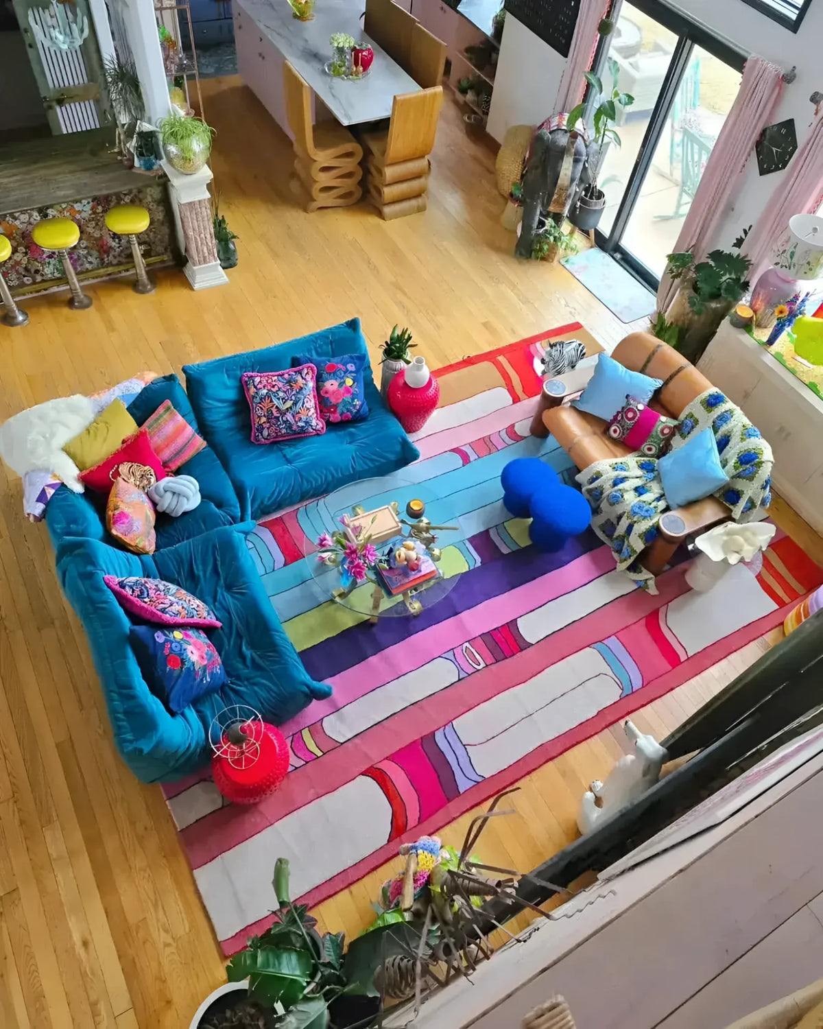 A vibrant and colorful Spectrum Stripe Hand Tufted Area Rug adorns a spacious living room with a contemporary design. The rug features bold, multicolored stripes that add a lively touch to the wooden floor. This wool rug, ideal for large living rooms, measures 8x10 and complements the eclectic, cozy seating arrangement, including a teal sofa and patterned cushions.