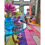 A vibrant Spectrum Stripe Hand Tufted Rug in a living room setting, showcasing its bold and colorful stripes. This contemporary wool rug adds a splash of color, complementing the eclectic decor with a glass coffee table and floral arrangements. Perfect for those seeking large living room rugs or an 8x10 area rug to enhance their space.