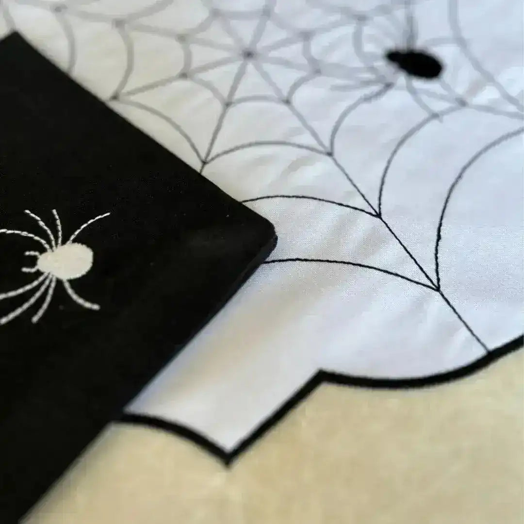 Spiderweb placemat detail, white scalloped mat with black embroidered web and spider at corner, black napkin at edge