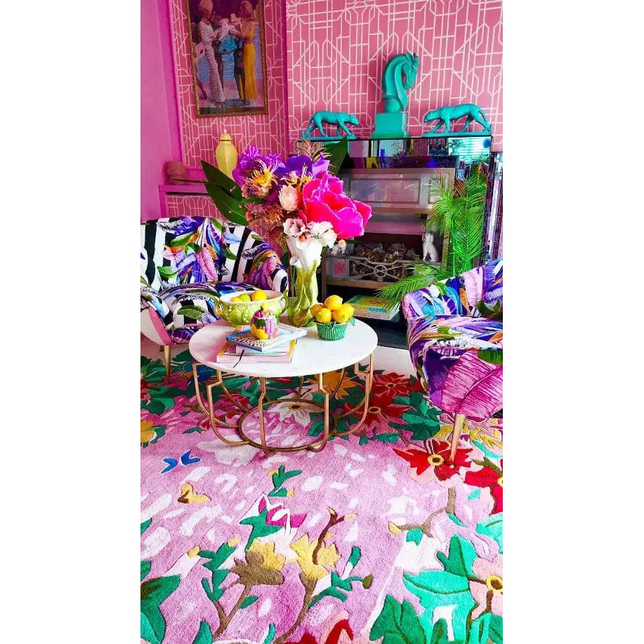 Vibrant living room featuring the Sunrise Bloom Hand Tufted Round Rug as a focal point. This large area rug showcases a colorful floral pattern, ideal for adding warmth and style to any living room. The wool rug's round shape complements the eclectic decor, with a white coffee table and colorful armchairs enhancing the cozy atmosphere.