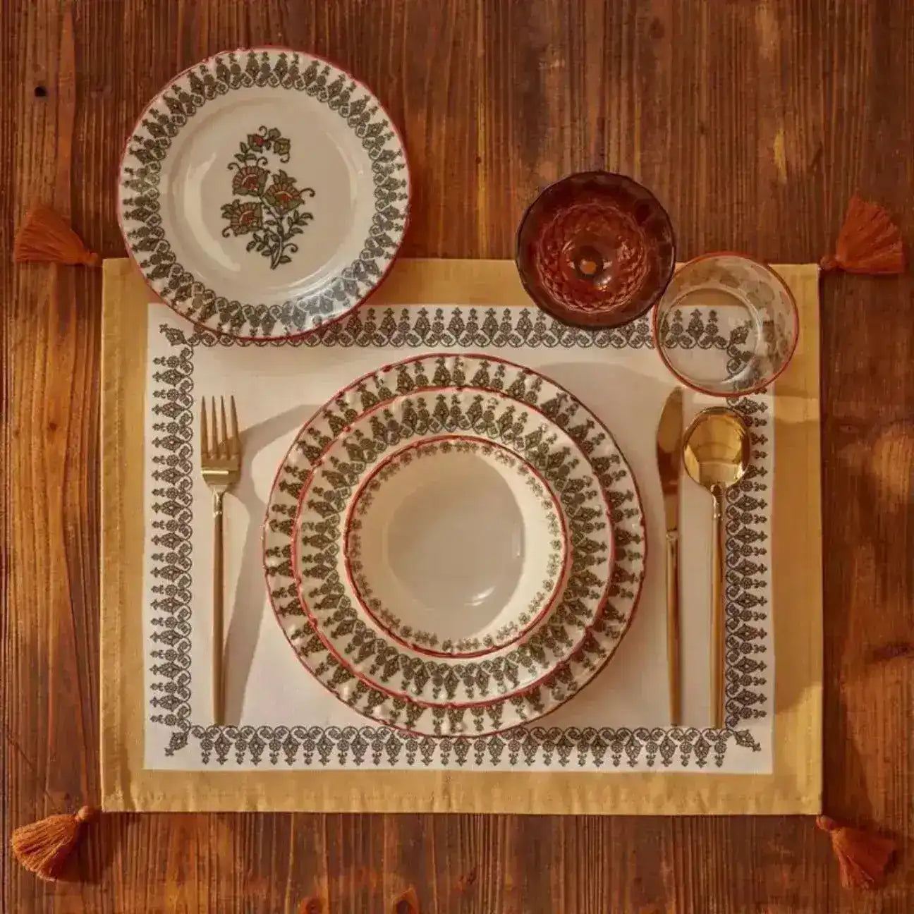 Tassel Trimmed placemat on wood table with stacked plates and gold cutlery, rust tassels at corners