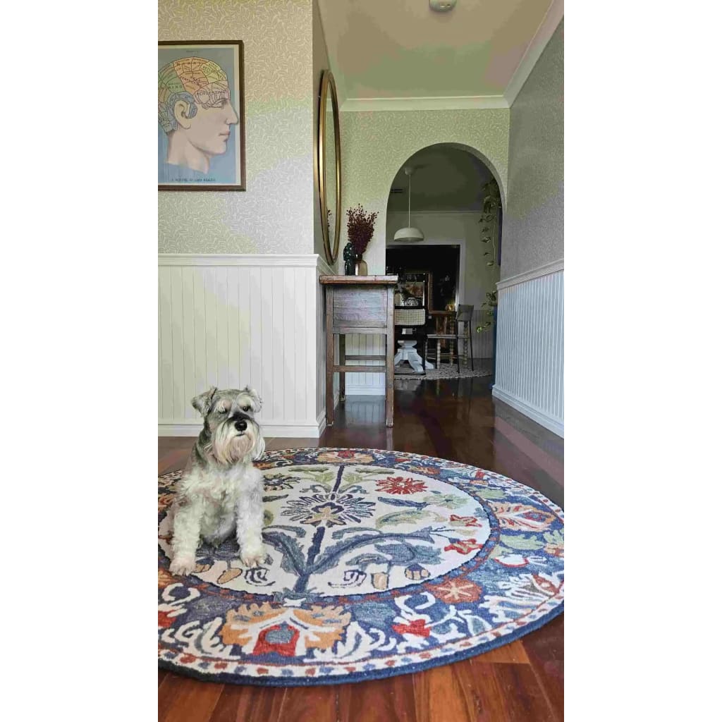 A charming dining room showcasing the Blossom Grace Hand Tufted Round Rug with vibrant floral patterns. This elegant wool rug adds a touch of sophistication to the hardwood floor, perfect for dining room area rugs. A small dog sits comfortably on the round rug, enhancing the cozy ambiance of the living space.