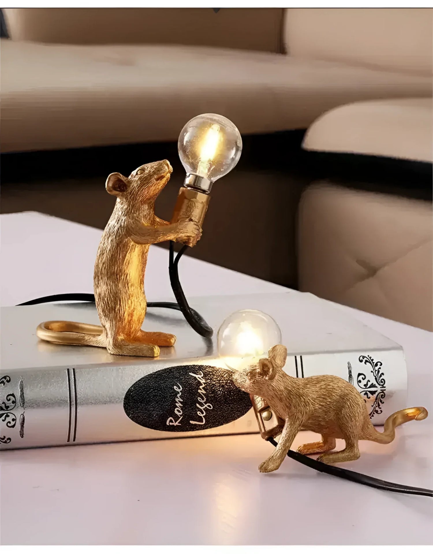 Two gold mice lamps on a book on coffee table, one standing, one crawling, both lit, sofa behind