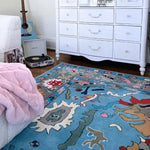 A vibrant Traditional Floral Hand Tufted Wool Rug in blue adorns a cozy living room. The area rug features an intricate floral design, adding a touch of elegance to the space. Perfect for those seeking rugs for living rooms, this wool rug complements the white dresser and warm wooden floor, creating a harmonious and inviting atmosphere.