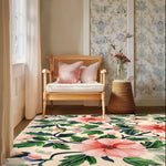 A beautiful hand-tufted area rug with a vibrant floral design graces a cozy living room. The wool rug, featuring lush green leaves and pink flowers, complements the soft tones of the wicker chair and pastel decor, making it an ideal choice for living room area rugs.