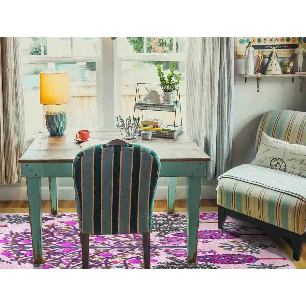 A vibrant Tree of Life Pink Mustard Hand Tufted Wool Rug adds a boho charm to this cozy living room. The large area rug, perfect for an 8x10 living room, features intricate patterns in pink and mustard tones, complementing the rustic wooden table and striped chair.