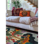 A vibrant Tropical Oasis Hand Tufted Area Rug in a living room setting. The wool rug features a colorful abstract design with tropical plant motifs, complementing the cozy white sofa adorned with decorative pillows. Ideal as a square area rug for living rooms, this 8x10 rug enhances the space with its exotic flair.