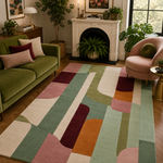 A large area rug with a mid-century modern abstract pattern in sage green, dusty pink, mustard orange, cream, and burgundy, styled in a cozy living room next to a green sofa and a pink armchair.